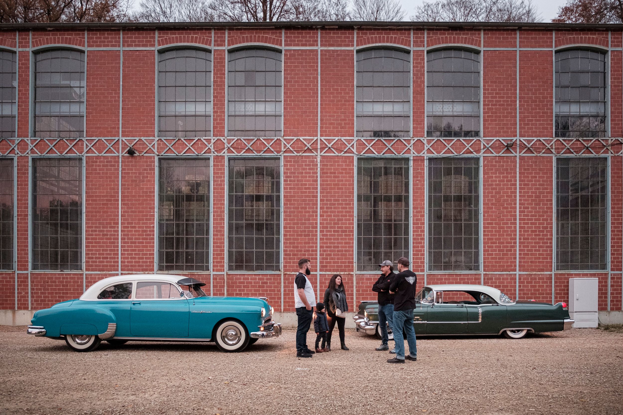 Zwei Classic-Cars stehen vor einem alten Industriebau, während ihre Besitzer sich austauschen